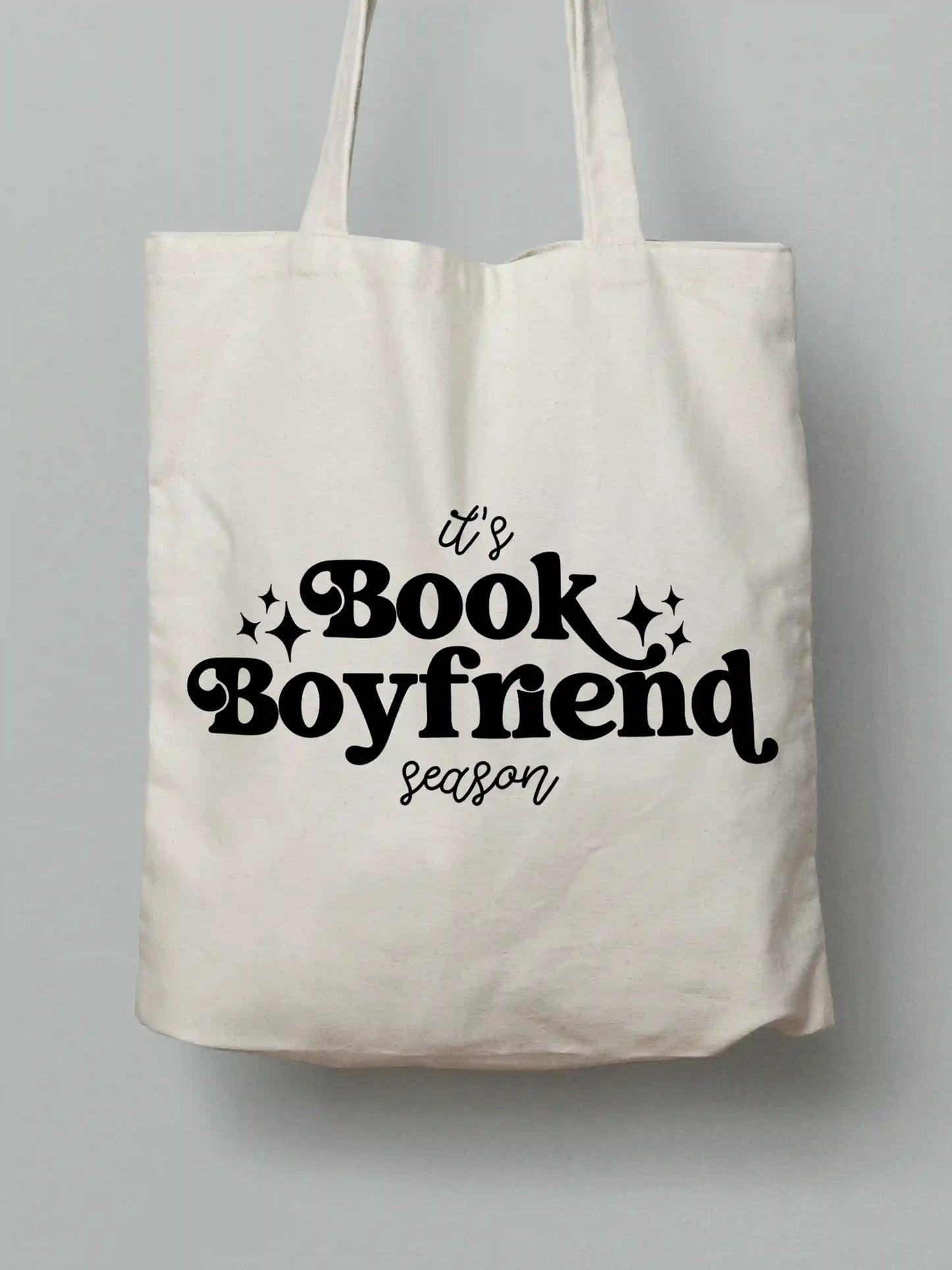 An white canvas tote bag with black text "It's Book Boyfriend season" and small black sparkle accents, shown against a solid gray background.