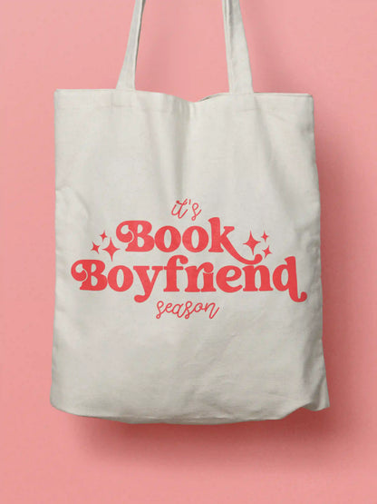 An white canvas tote bag with peach text "It's Book Boyfriend season" and small peach sparkle accents, shown against a solid peach background.