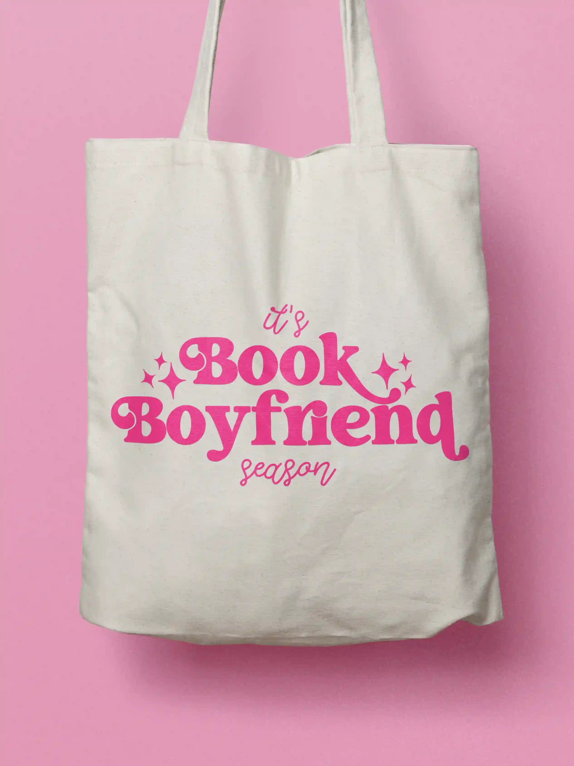 An white canvas tote bag with pink text "It's Book Boyfriend season" and small pink sparkle accents, shown against a solid pink background.