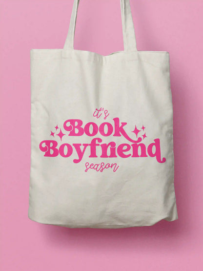 An white canvas tote bag with pink text "It's Book Boyfriend season" and small pink sparkle accents, shown against a solid pink background.