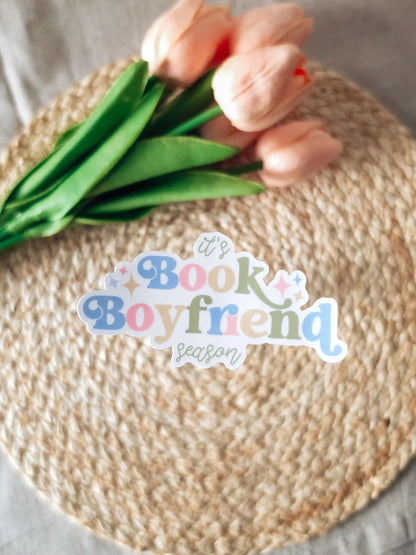 A die-cut sticker reading "it's Book Boyfriend season" in blue, pink, and green lettering with sparkle details, placed on a woven rattan mat with peach roses and green leaves resting in the upper corner.