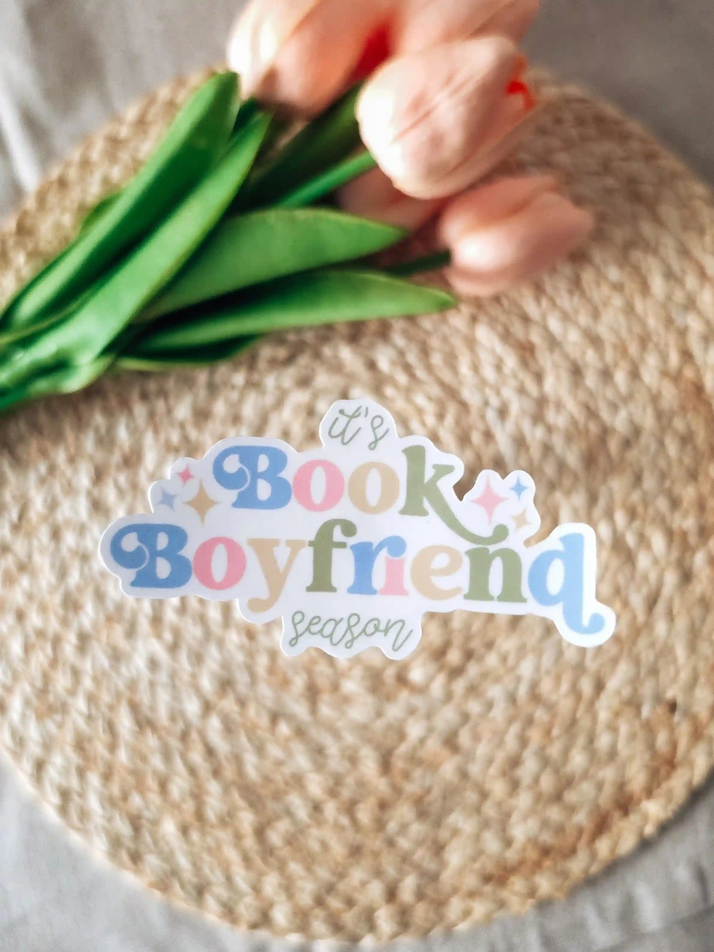 A die-cut sticker reading "it's Book Boyfriend season" in blue, pink, and green lettering with sparkle details, placed on a woven rattan mat with peach roses and green leaves resting in the upper corner.