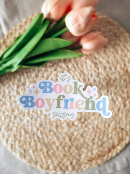 A die-cut sticker reading "it's Book Boyfriend season" in blue, pink, and green lettering with sparkle details, placed on a woven rattan mat with peach roses and green leaves resting in the upper corner.