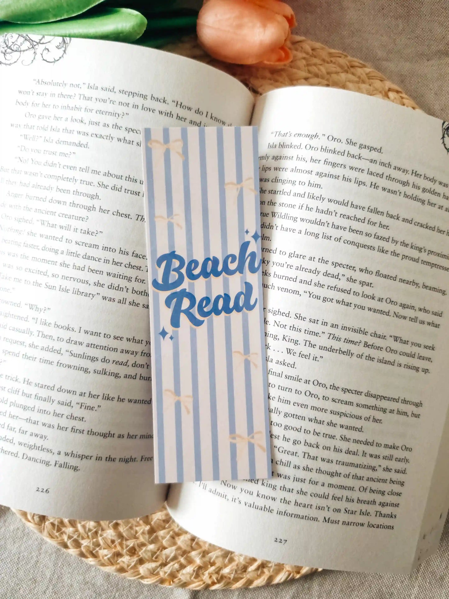 A striped paper bookmark with blue "Beach Read" lettering and small stars, placed across an open book on a woven placemat with tulips in the background.