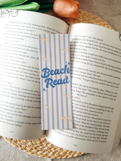 A striped paper bookmark with blue "Beach Read" lettering and small stars, placed across an open book on a woven placemat with tulips in the background.
