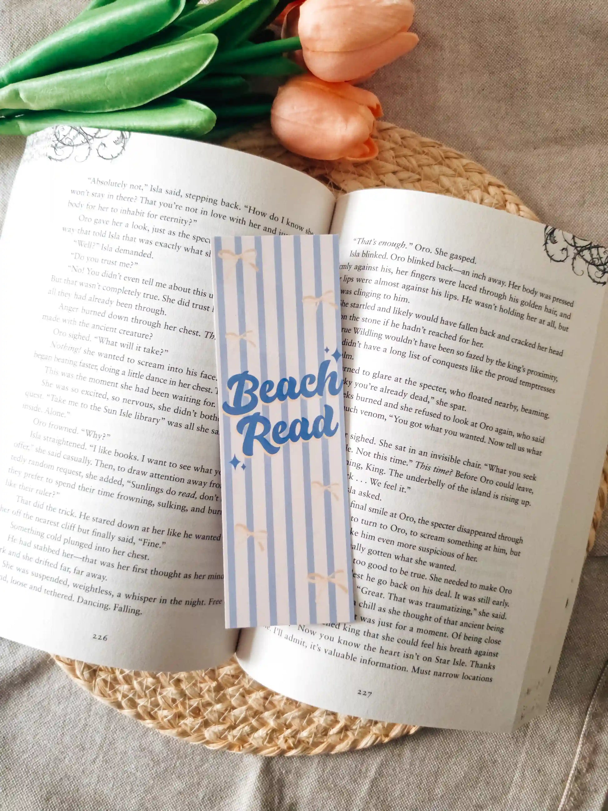 A striped paper bookmark with blue "Beach Read" lettering and small stars, placed across an open book on a woven placemat with tulips in the background.
