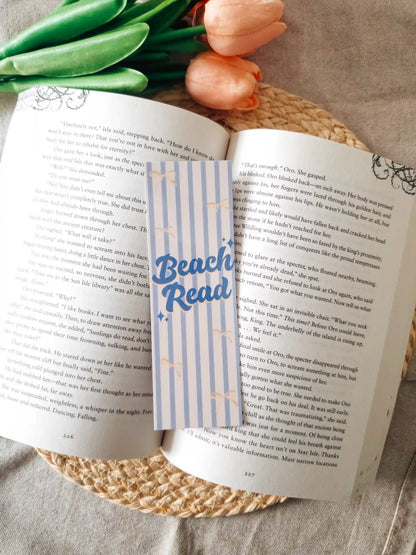 A striped paper bookmark with blue "Beach Read" lettering and small stars, placed across an open book on a woven placemat with tulips in the background.