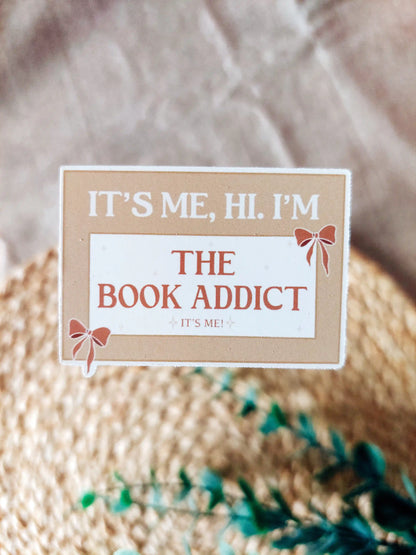 Beige vinyl sticker with the phrase "It’s me, hi. I’m the book addict", photographed on a woven neutral background.