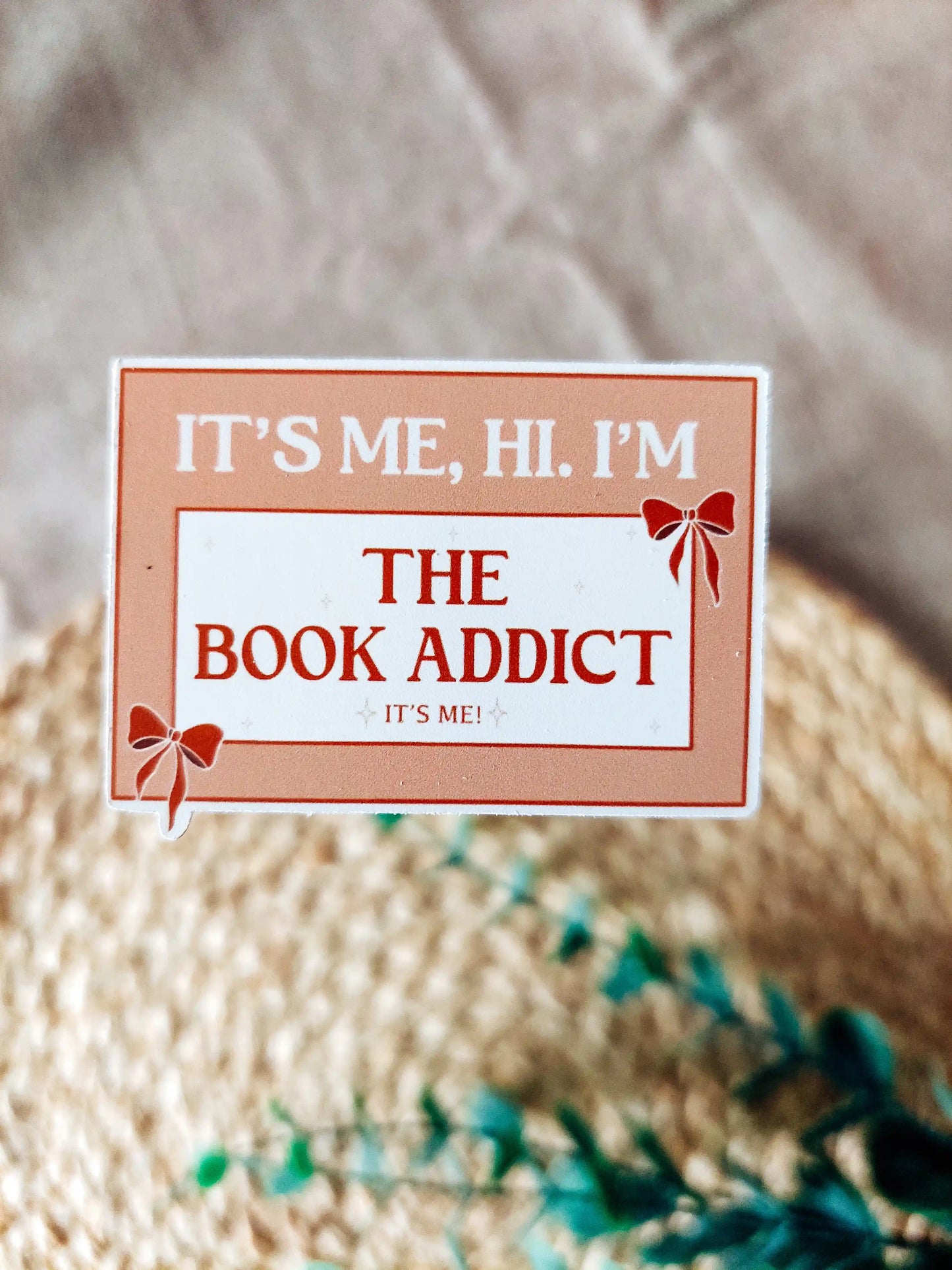 Orange vinyl sticker with the phrase "It’s me, hi. I’m the book addict", photographed on a woven neutral background.
