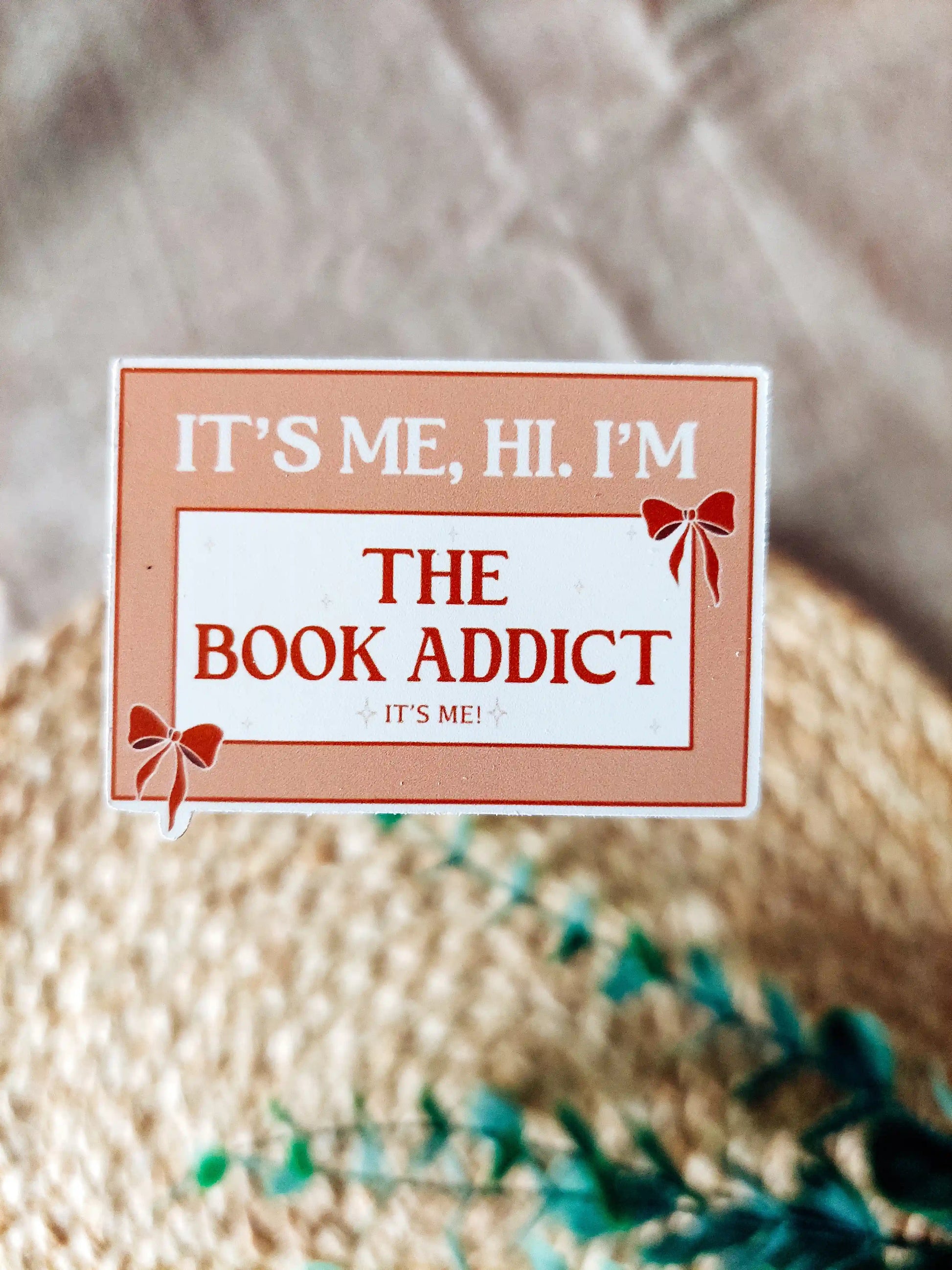 Orange vinyl sticker with the phrase "It’s me, hi. I’m the book addict", photographed on a woven neutral background.