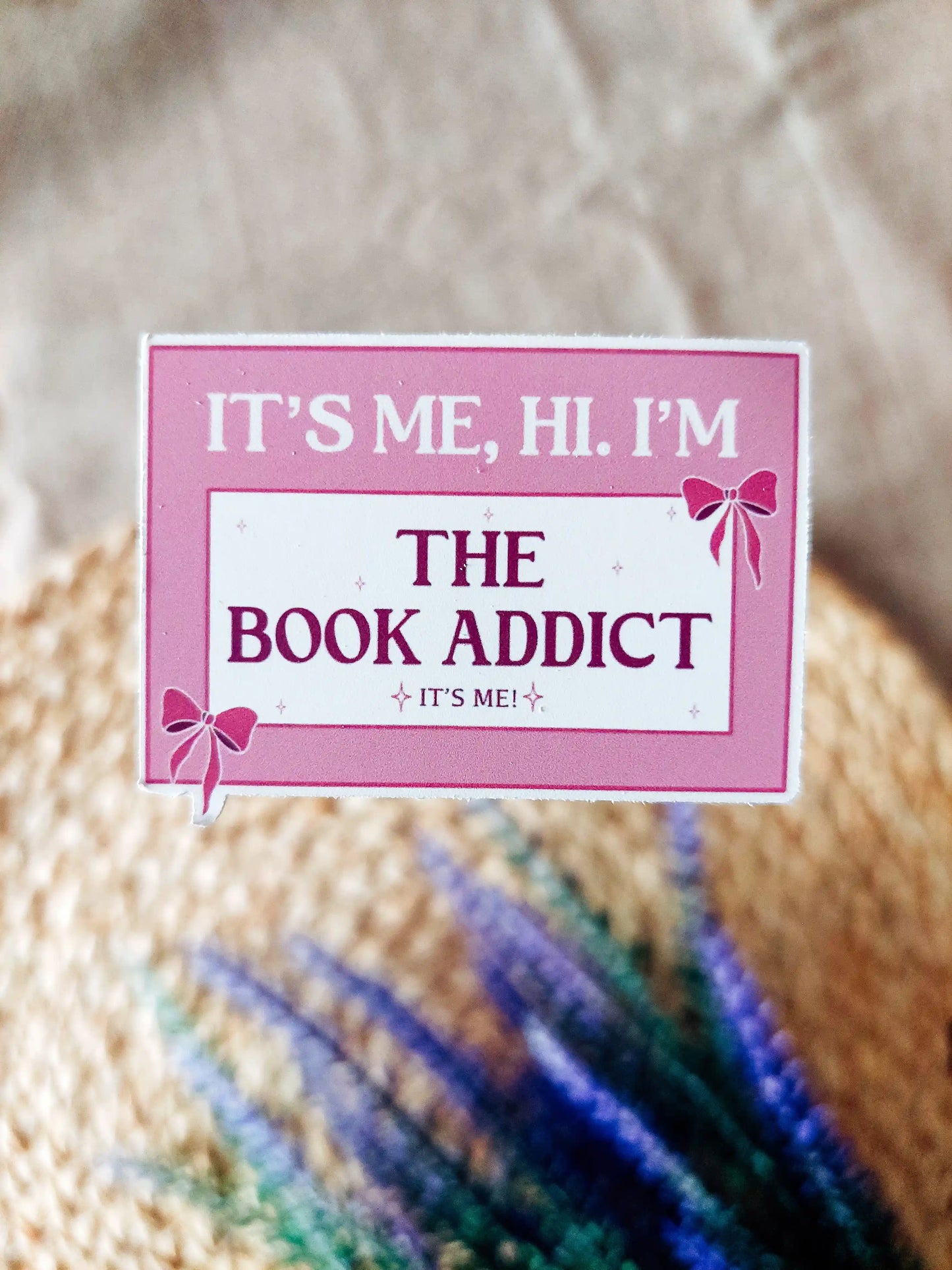 Pink vinyl sticker with the phrase "It’s me, hi. I’m the book addict", photographed on a woven neutral background.