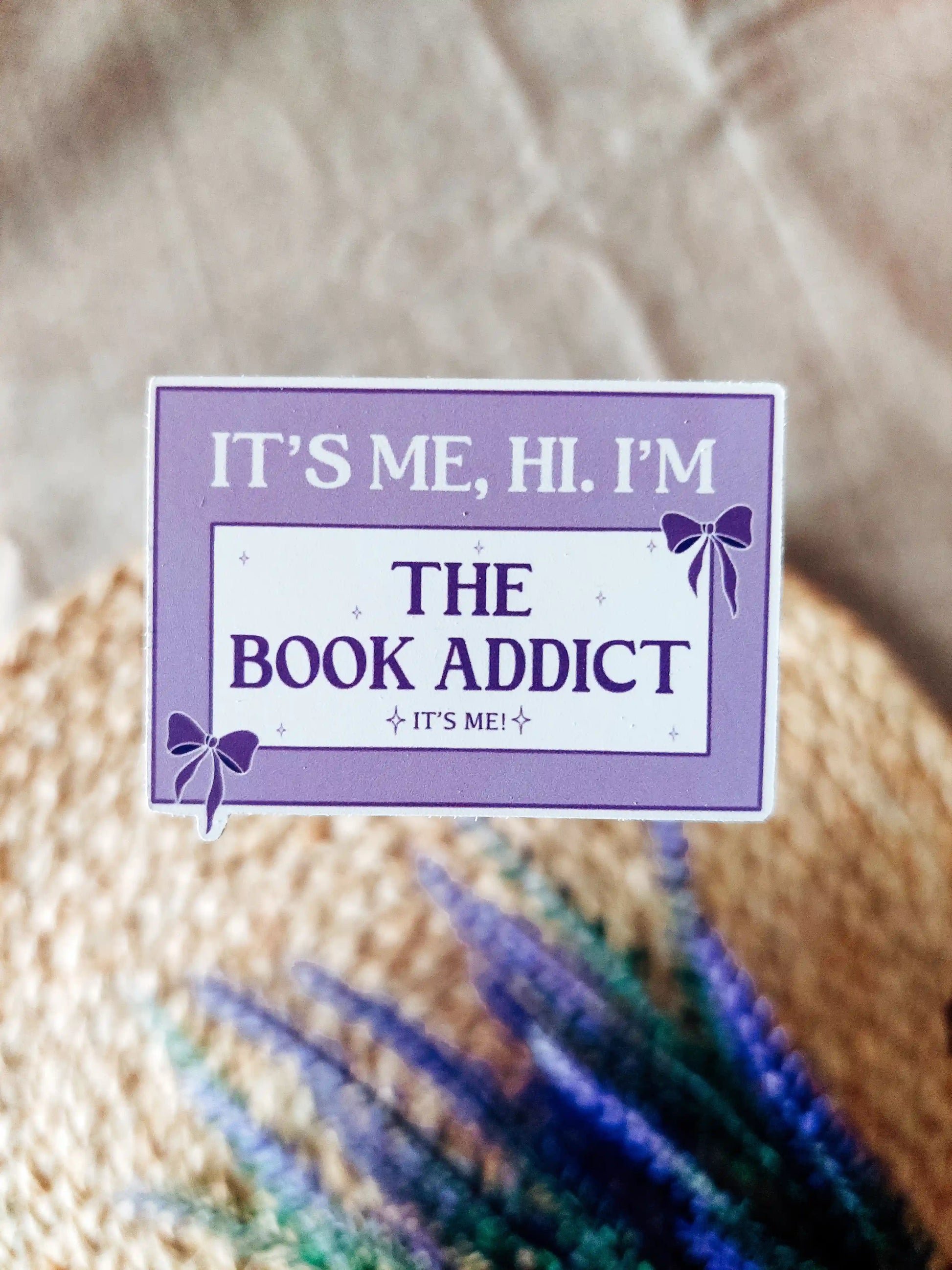 Purple vinyl sticker with the phrase "It’s me, hi. I’m the book addict", photographed on a woven neutral background.