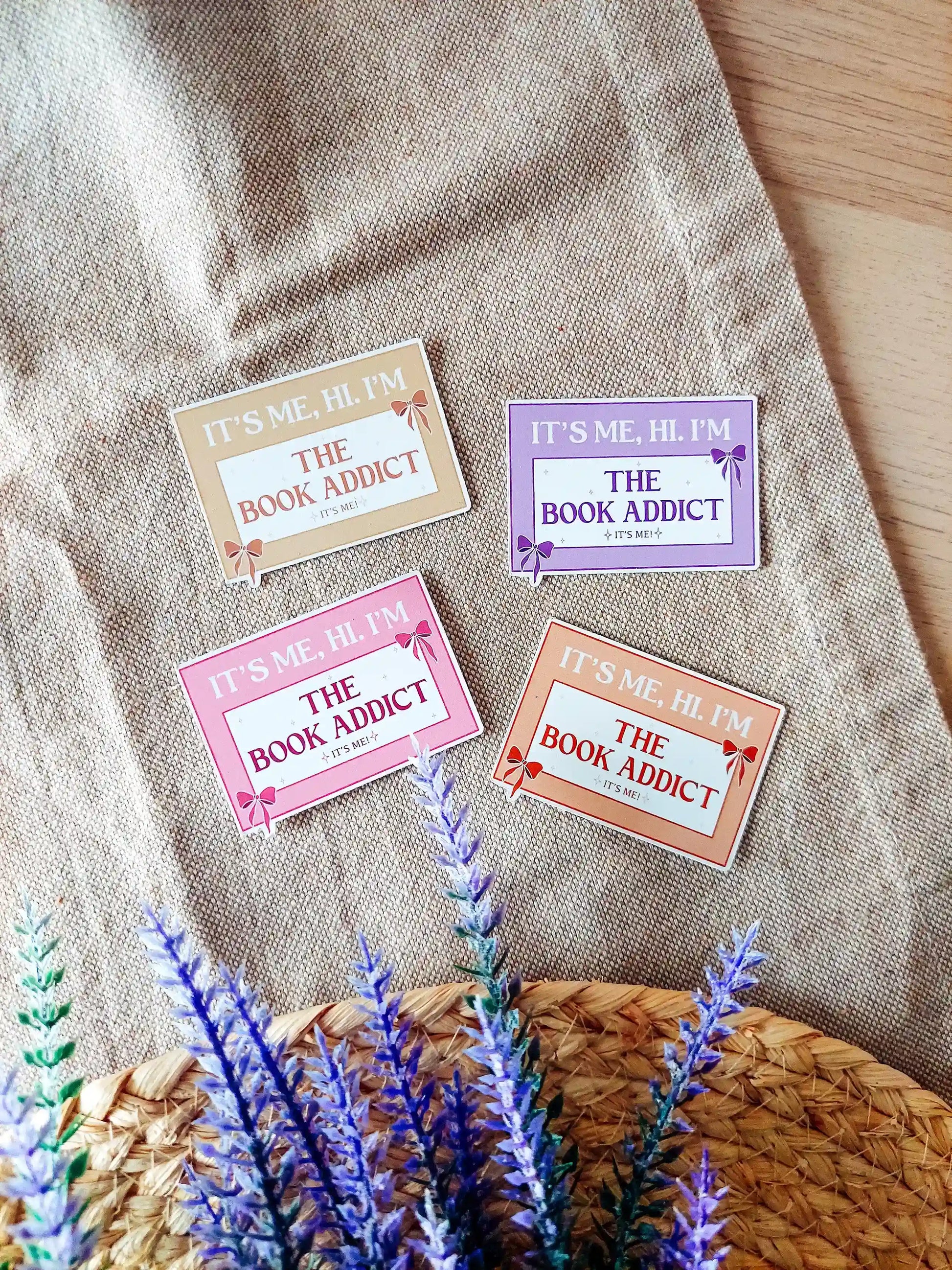 Vinyl stickers in four color variants with the phrase "It’s me, hi. I’m the Book Addict", displayed on a neutral textured background.