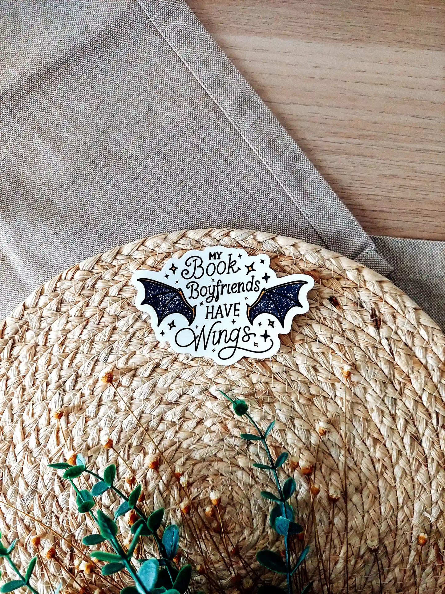Vinyl sticker with the text "My book boyfriends have wings" with black glittery bat wings and star accents, photographed on a woven neutral background.