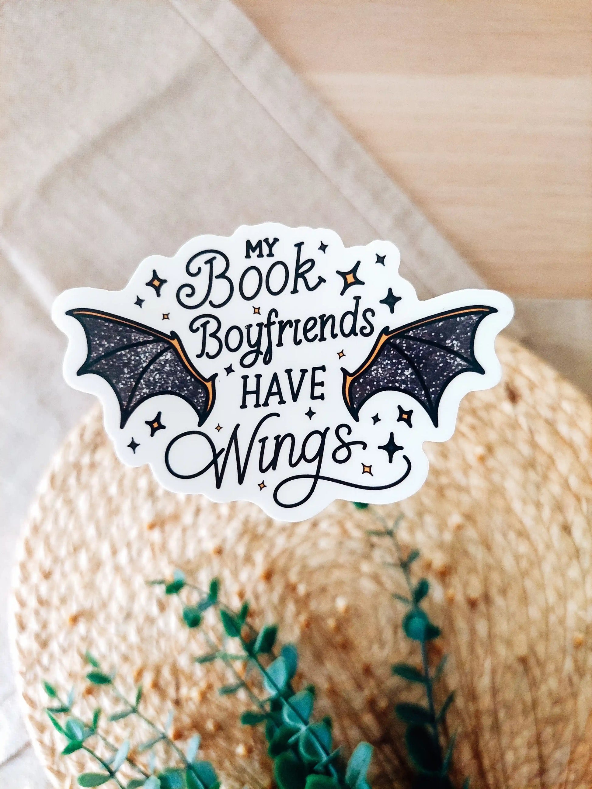 Vinyl sticker with the text "My book boyfriends have wings" with black glittery bat wings and star accents, photographed on a woven neutral background.