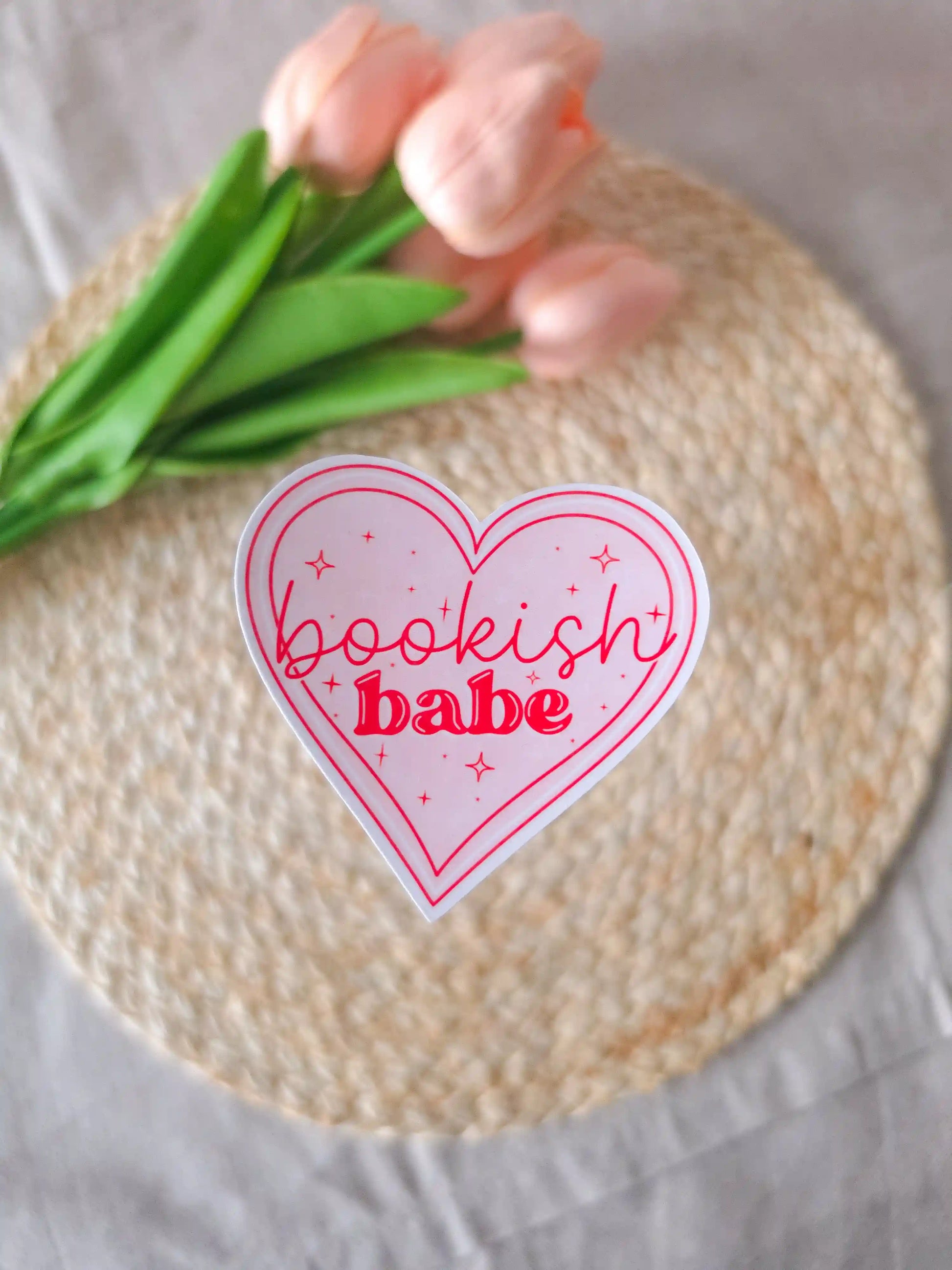 A heart-shaped die-cut sticker with a light pink background, red double outline border, sparkle details, and the text "Bookish Babe" in red lettering, placed on a woven rattan mat. Peach tulips with green leaves in the upper corner.