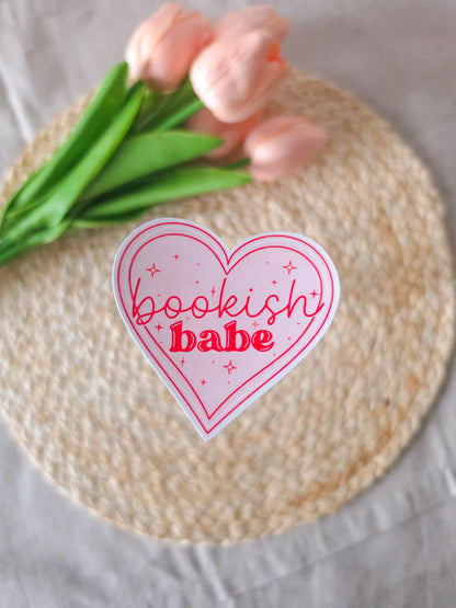 A heart-shaped die-cut sticker with a light pink background, red double outline border, sparkle details, and the text "Bookish Babe" in red lettering, placed on a woven rattan mat. Peach tulips with green leaves in the upper corner.