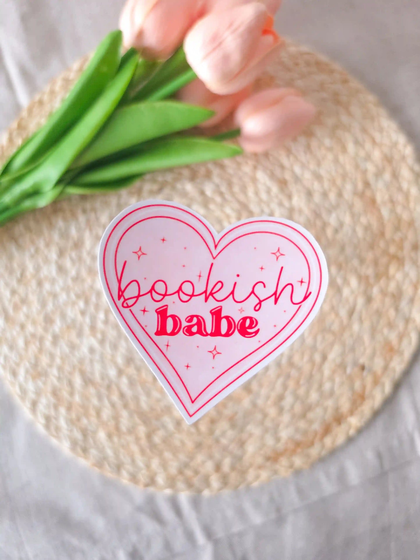 A heart-shaped die-cut sticker with a light pink background, red double outline border, sparkle details, and the text "Bookish Babe" in red lettering, placed on a woven rattan mat. Peach tulips with green leaves in the upper corner.