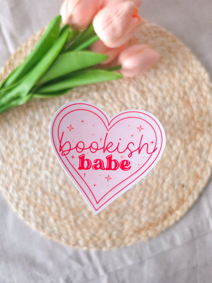 A heart-shaped die-cut sticker with a light pink background, red double outline border, sparkle details, and the text "Bookish Babe" in red lettering, placed on a woven rattan mat. Peach tulips with green leaves in the upper corner.