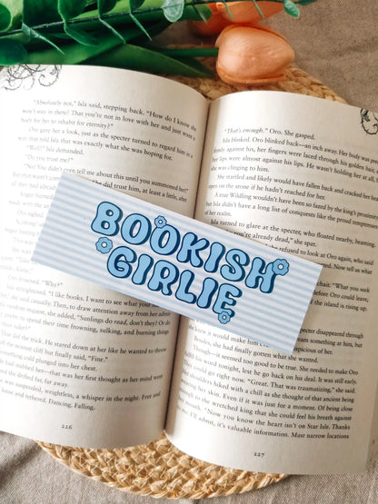 A bookmark with the words 
"BOOKISH GIRLIE" in blue bubble letters, laid over an open book. The scene includes pink fake roses and green leaves at the top, set on a woven jute surface.