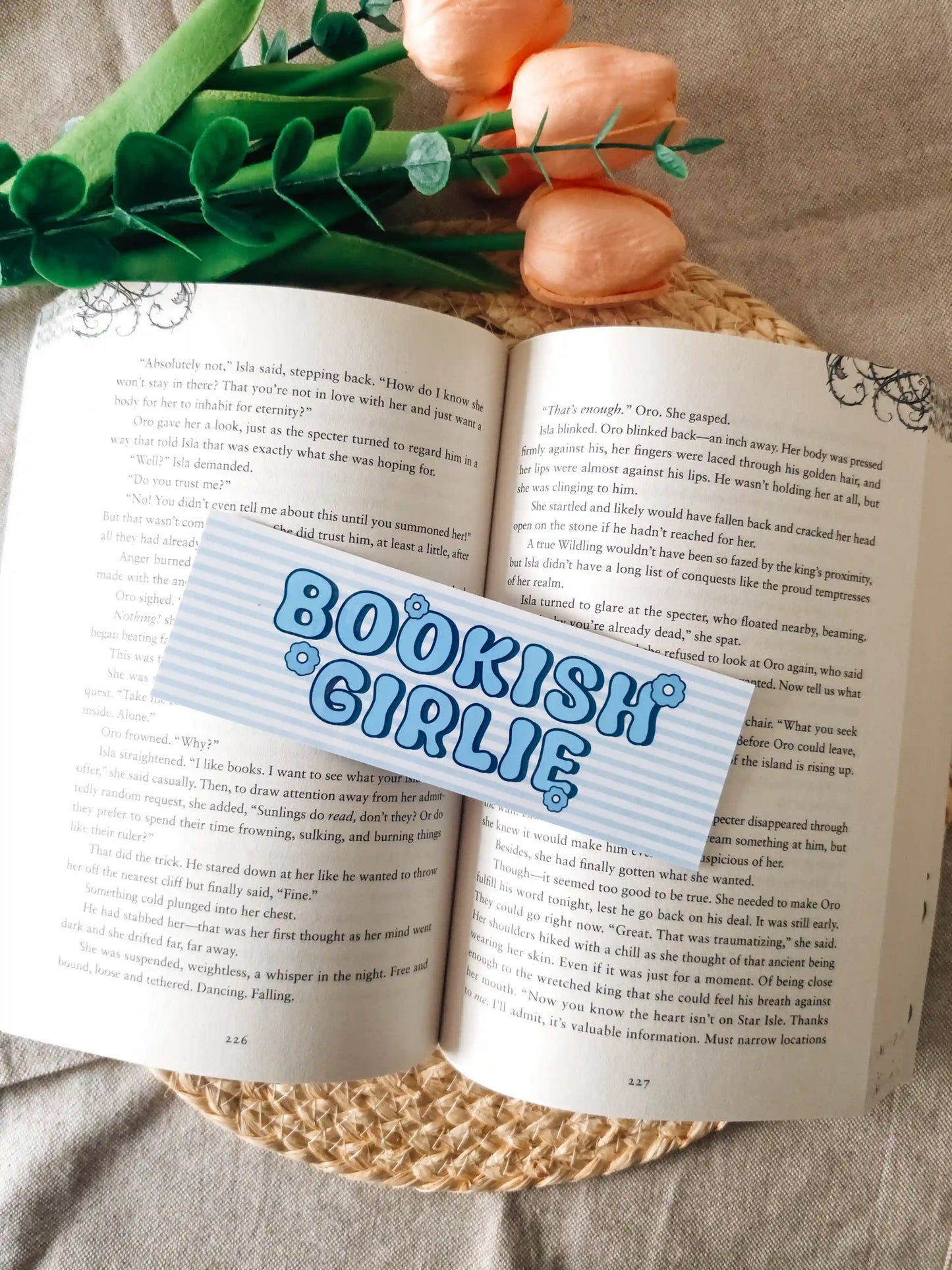 A bookmark with the words 
"BOOKISH GIRLIE" in blue bubble letters, laid over an open book. The scene includes pink fake roses and green leaves at the top, set on a woven jute surface.