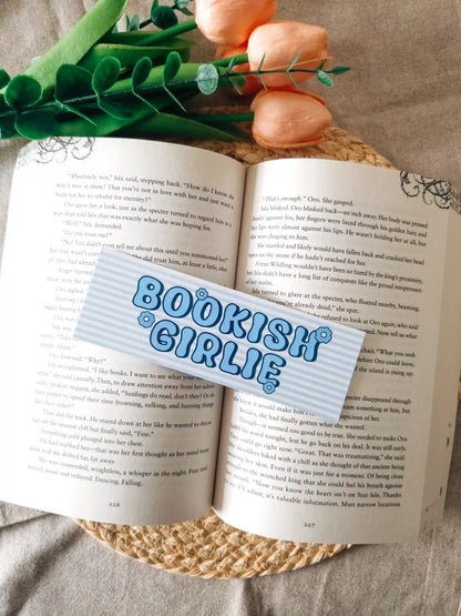 A bookmark with the words 
"BOOKISH GIRLIE" in blue bubble letters, laid over an open book. The scene includes pink fake roses and green leaves at the top, set on a woven jute surface.