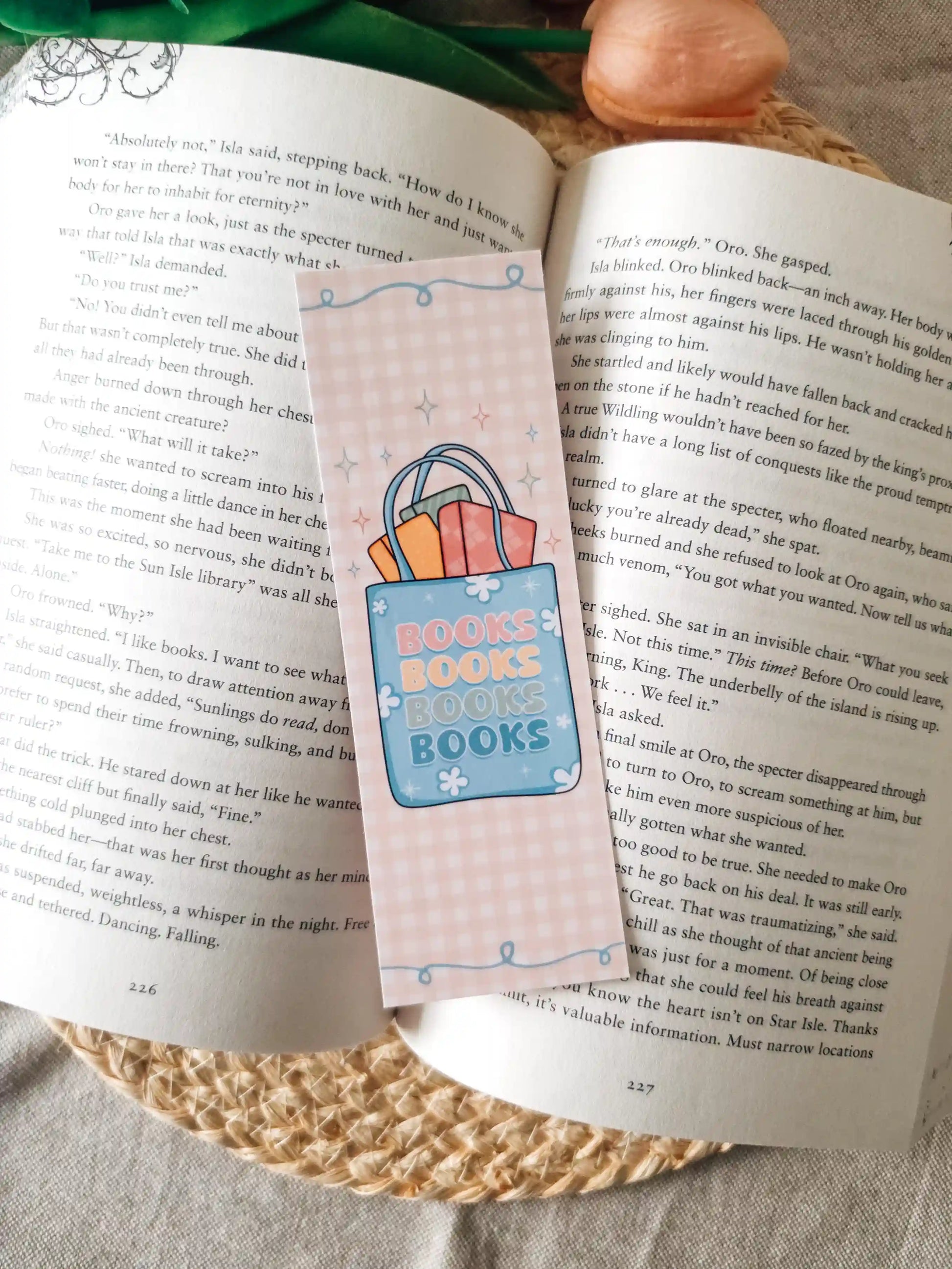 A pastel pink paper bookmark with a overflowing tote bag illustration with the text "books books boks books" placed on an open book, styled with green leaves and peach flowers on a woven mat.