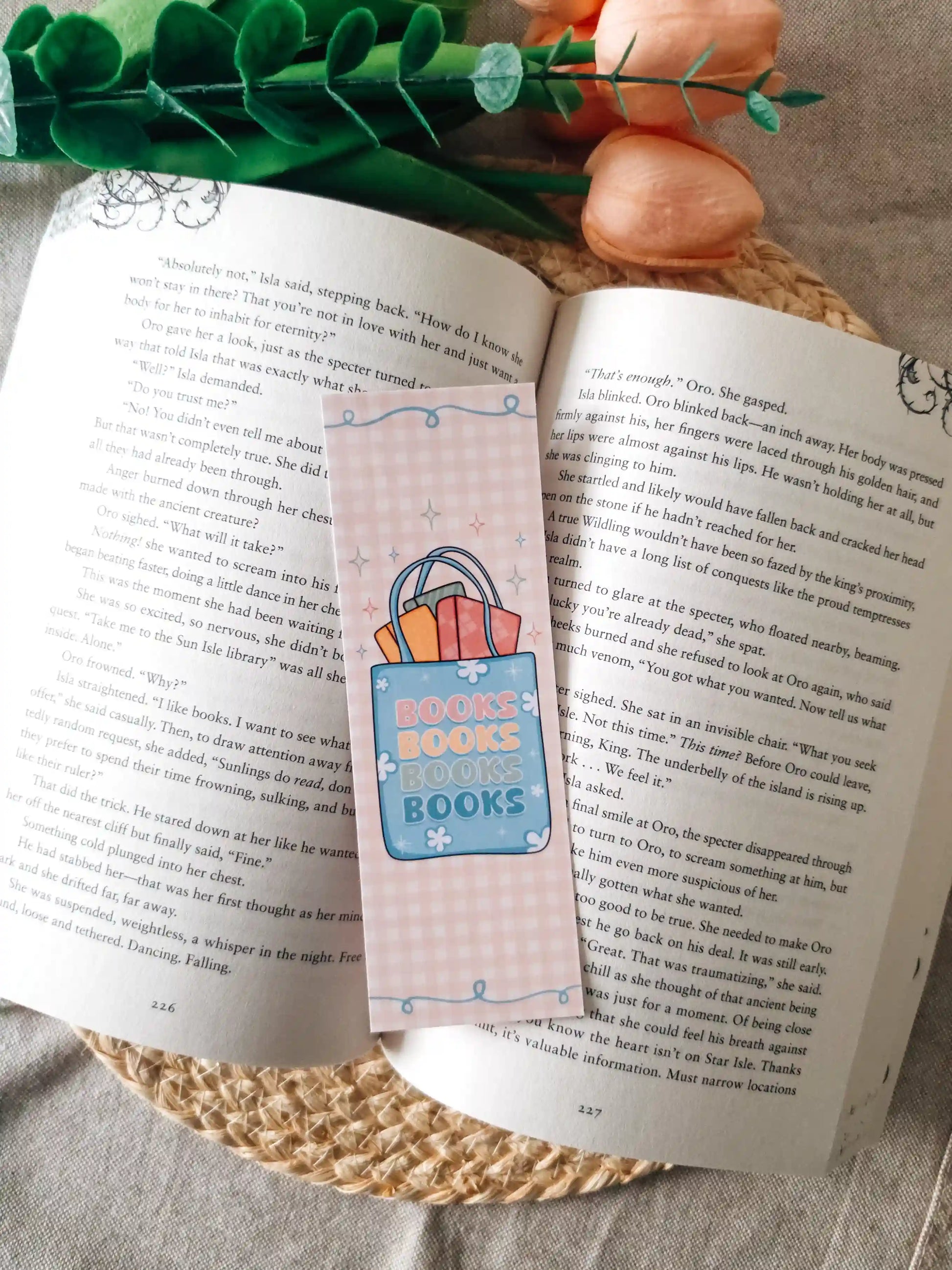 A pastel pink paper bookmark with a overflowing tote bag illustration with the text "books books boks books" placed on an open book, styled with green leaves and peach flowers on a woven mat.