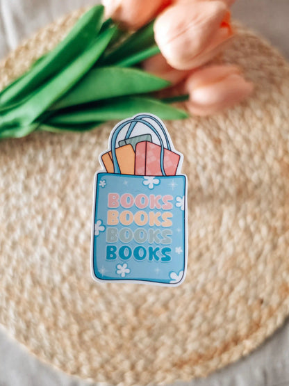 A die-cut sticker featuring a blue tote bag filled with colorful books and the text "Books Books Books Books" in pink, peach, and teal lettering, placed on a woven rattan mat. In the background, peach tulips with green leaves.