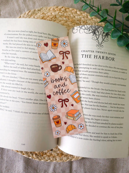 A laminated bookmark on a brown and blush checkered background featuring the text "books and coffee" in dark cursive lettering at the centre, surrounded by illustrated coffee cups with hearts, croissants, open books, stacked books, red bows, white daisies, and small hearts, resting on an open book on a woven rattan mat, with green eucalyptus sprigs in the upper right corner.