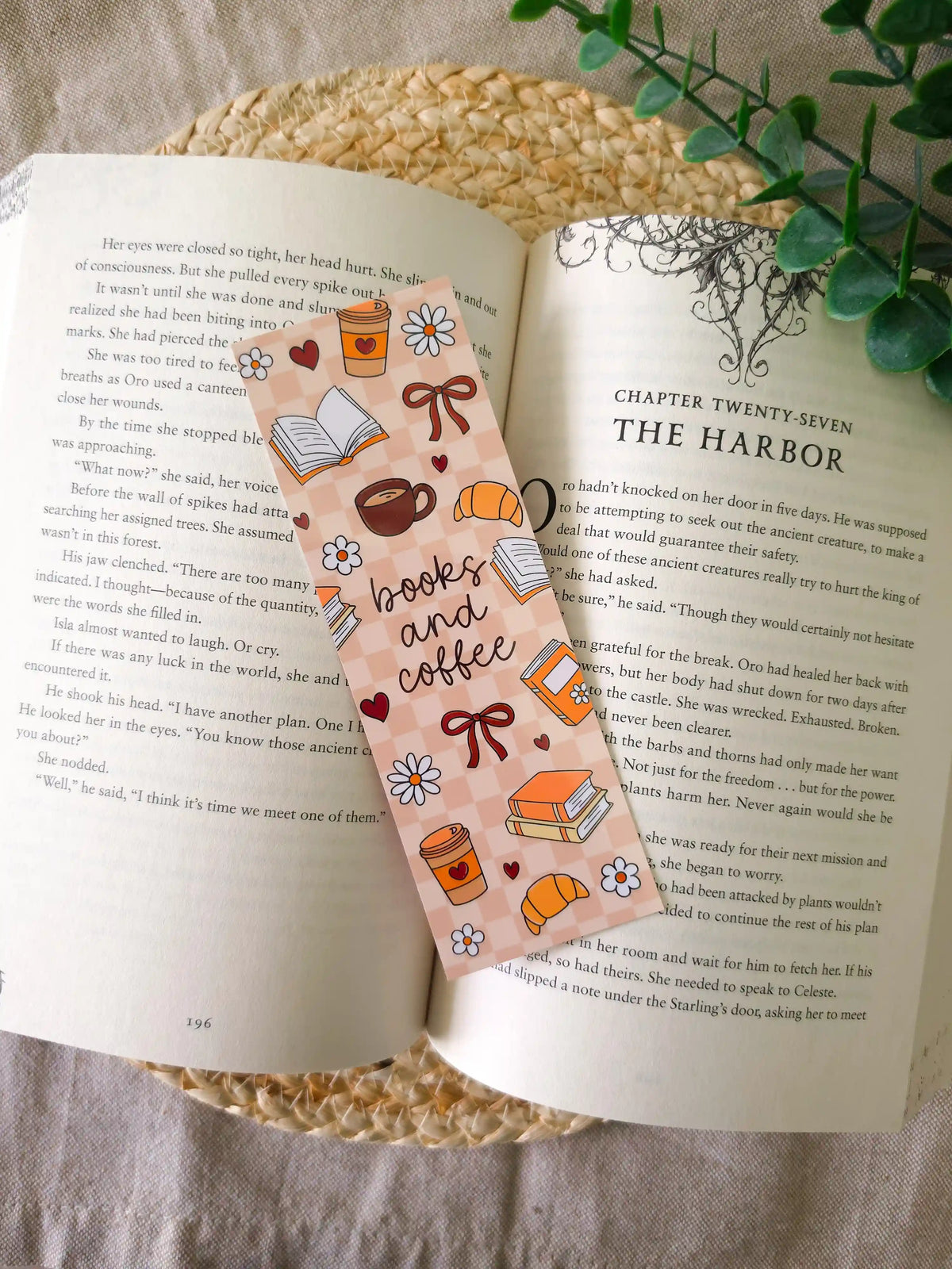 A laminated bookmark on a brown and blush checkered background featuring the text "books and coffee" in dark cursive lettering at the centre, surrounded by illustrated coffee cups with hearts, croissants, open books, stacked books, red bows, white daisies, and small hearts, resting on an open book on a woven rattan mat, with green eucalyptus sprigs in the upper right corner.