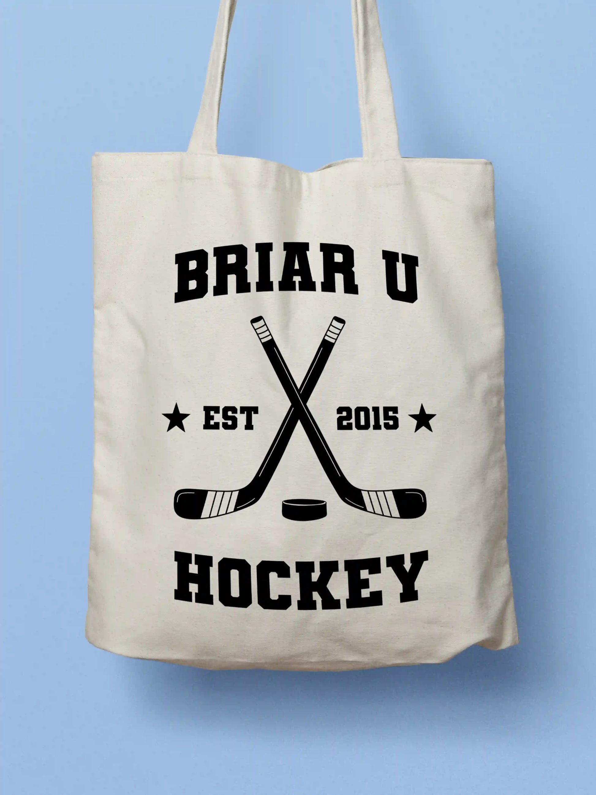 A white canvas tote bag featuring a "Briar U Hockey Est. 2015" design with crossed hockey sticks and a puck in black collegiate-style lettering, photographed against a light blue background.