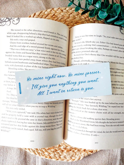 Bookmark inspired by Bridgerton featuring the quote "Be mine right now. Be mine forever. I'll give you anything you want. All I want in return is you", displayed on an open book over a woven mat with decorative plants. 