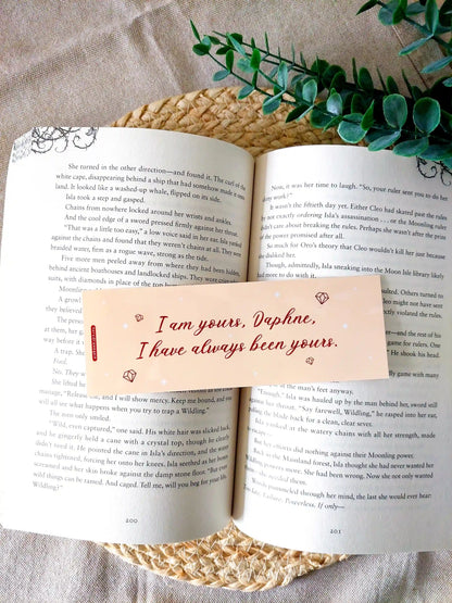 Bookmark inspired by Bridgerton featuring the quote "I am yours, Daphne. I have always been yours", displayed on an open book over a woven mat with decorative plants. 