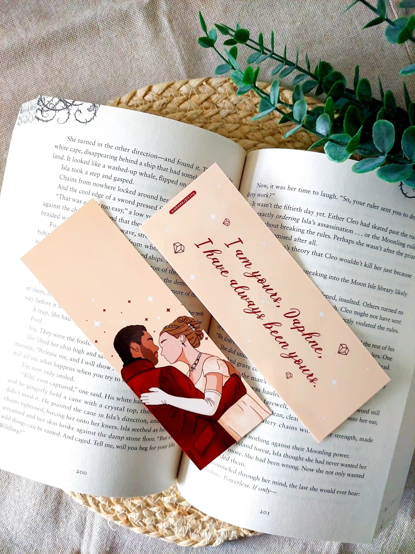 Bookmark inspired by Bridgerton featuring an illustration of Daphne and Simon on the front and the quote "I am yours, Daphne. I have always been yours" on the back, displayed on an open book over a woven mat with decorative plants. 