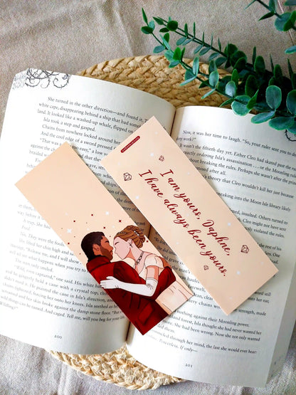 Bookmark inspired by Bridgerton featuring an illustration of Daphne and Simon on the front and the quote "I am yours, Daphne. I have always been yours" on the back, displayed on an open book over a woven mat with decorative plants. 