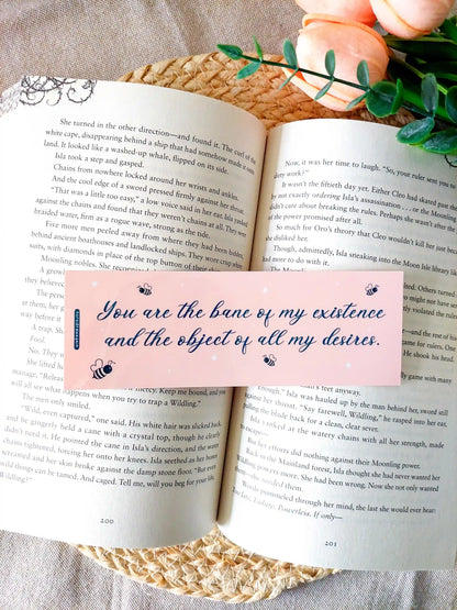 Bookmark inspired by Bridgerton featuring the quote "You are the bane of my existence and the object of all my desires", displayed on an open book over a woven mat with decorative plants. 
