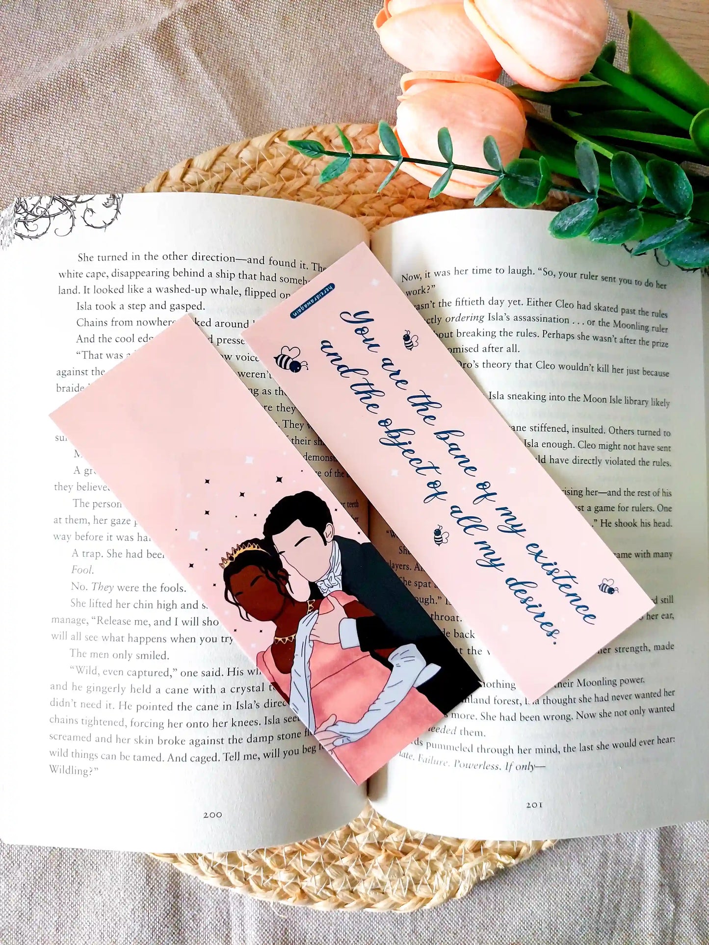 Bookmark inspired by Bridgerton featuring an illustration of Kate and Anthony on the front and the quote "You are the bane of my existence and the object of all my desires" on the back, displayed on an open book over a woven mat with decorative plants. 