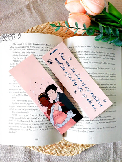 Bookmark inspired by Bridgerton featuring an illustration of Kate and Anthony on the front and the quote "You are the bane of my existence and the object of all my desires" on the back, displayed on an open book over a woven mat with decorative plants. 
