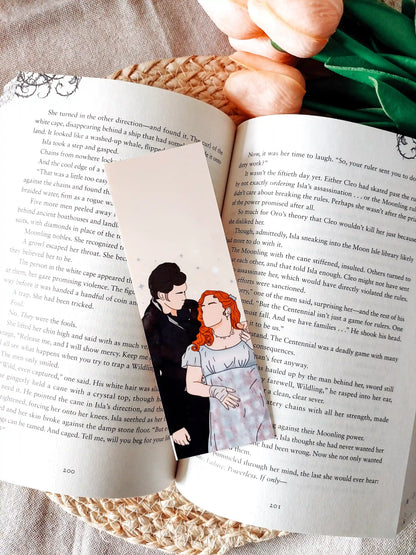 Bookmark inspired by Bridgerton featuring an illustration of Penelope and Colin, displayed on an open book over a woven mat with decorative plants. 