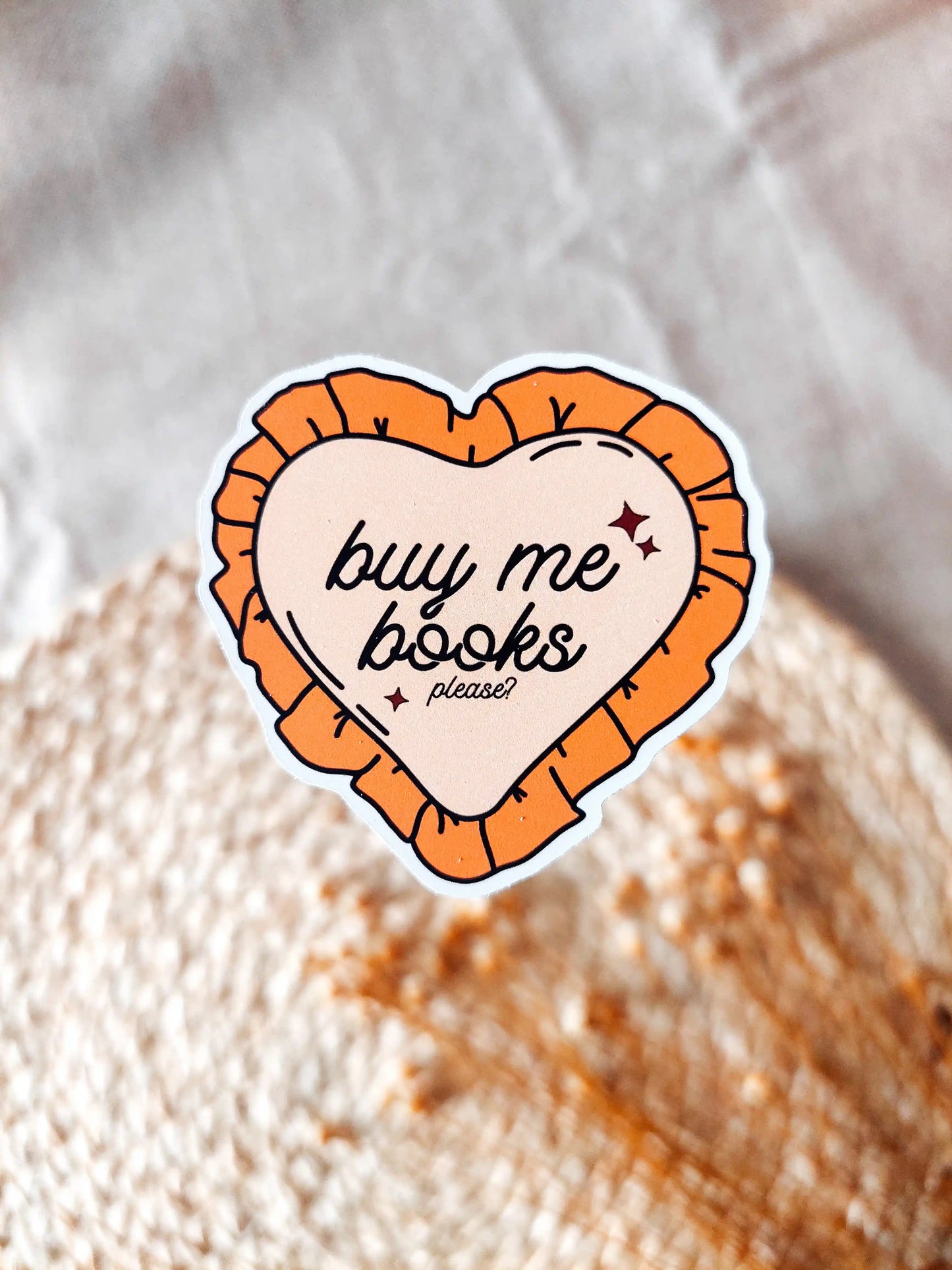Orange heart-shaped vinyl sticker reading "Buy me books, please", photographed on a woven neutral background.