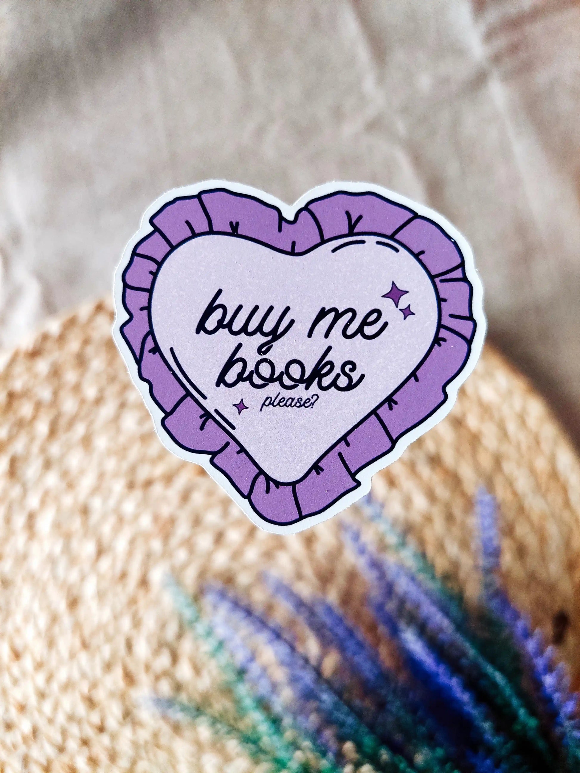 Purple heart-shaped vinyl sticker reading "Buy me books, please", shown on a textured woven surface.