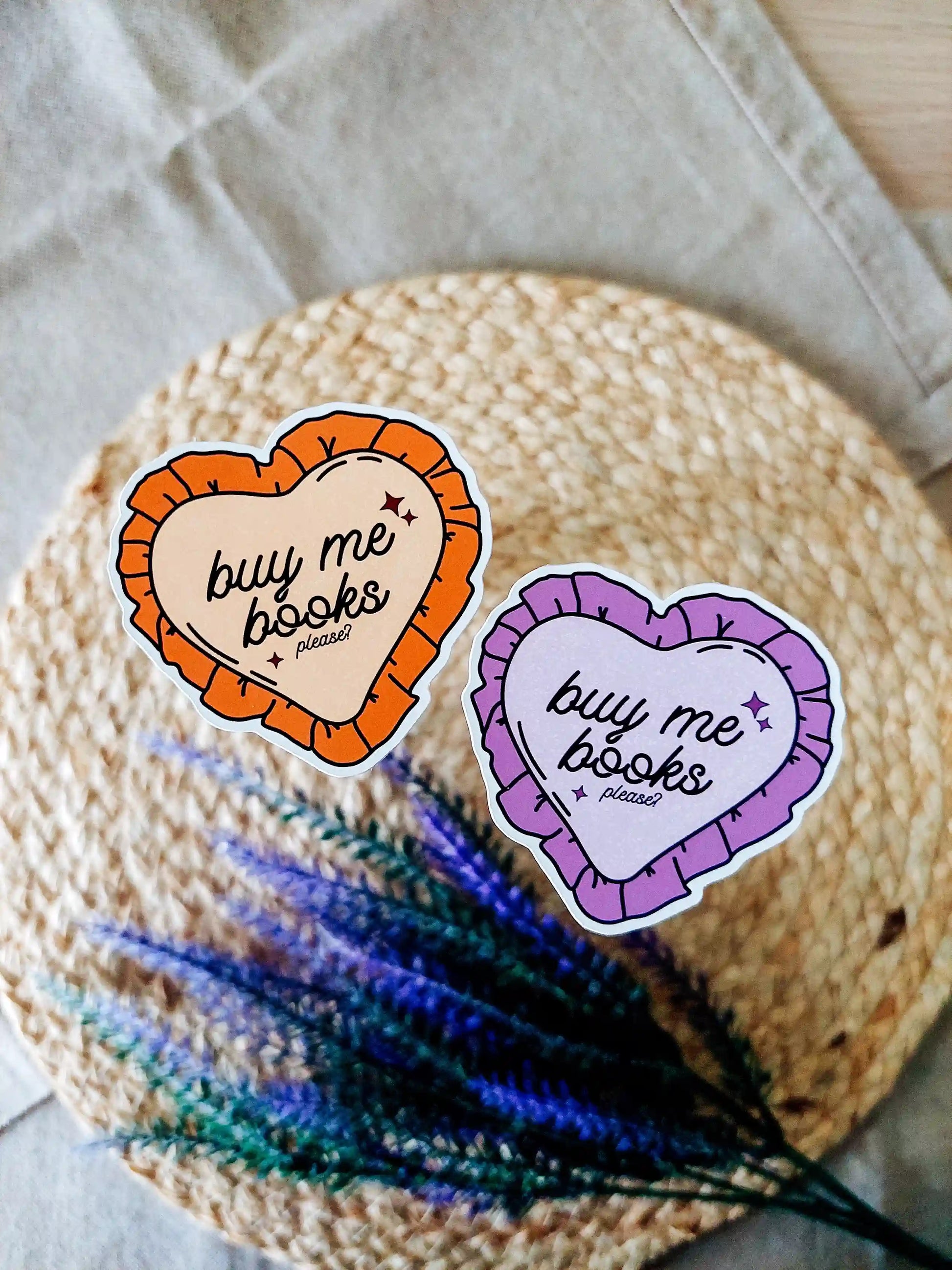 Two heart-shaped vinyl stickers reading "Buy me books, please" in purple and orange color variants, photographed on a woven neutral background.
