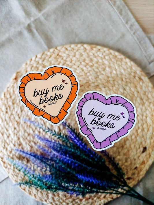 Two heart-shaped vinyl stickers reading "Buy me books, please" in purple and orange color variants, photographed on a woven neutral background.