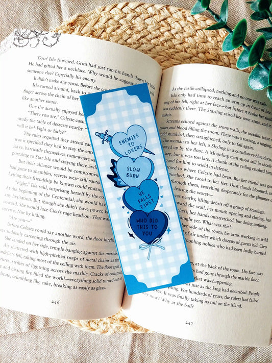 Candy hearts tropes bookmark placed on an open book, featuring Enemies to Lovers, Slow Burn, He Falls First and Who Did This To You in shades of blue.