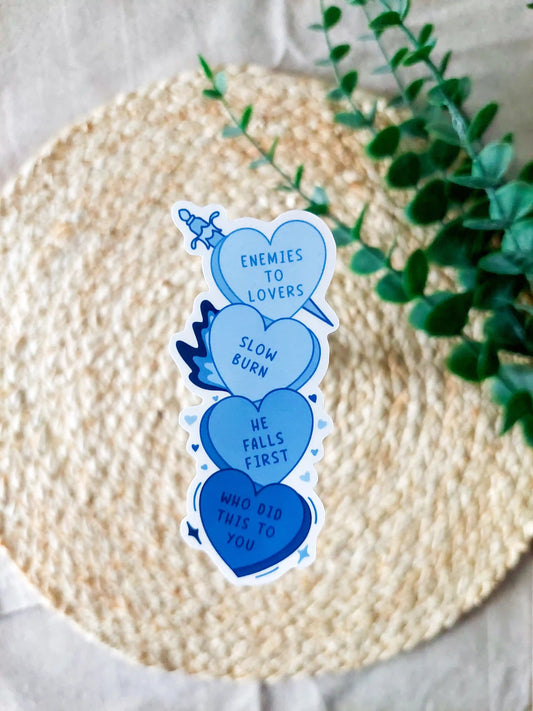 A pastel blue sticker inspired by candy hearts featuring romance book tropes including "Enemies to Lovers", "Slow Burn" and "He Falls First". The sticker is displayed on a woven beige surface with soft greenery in the background.