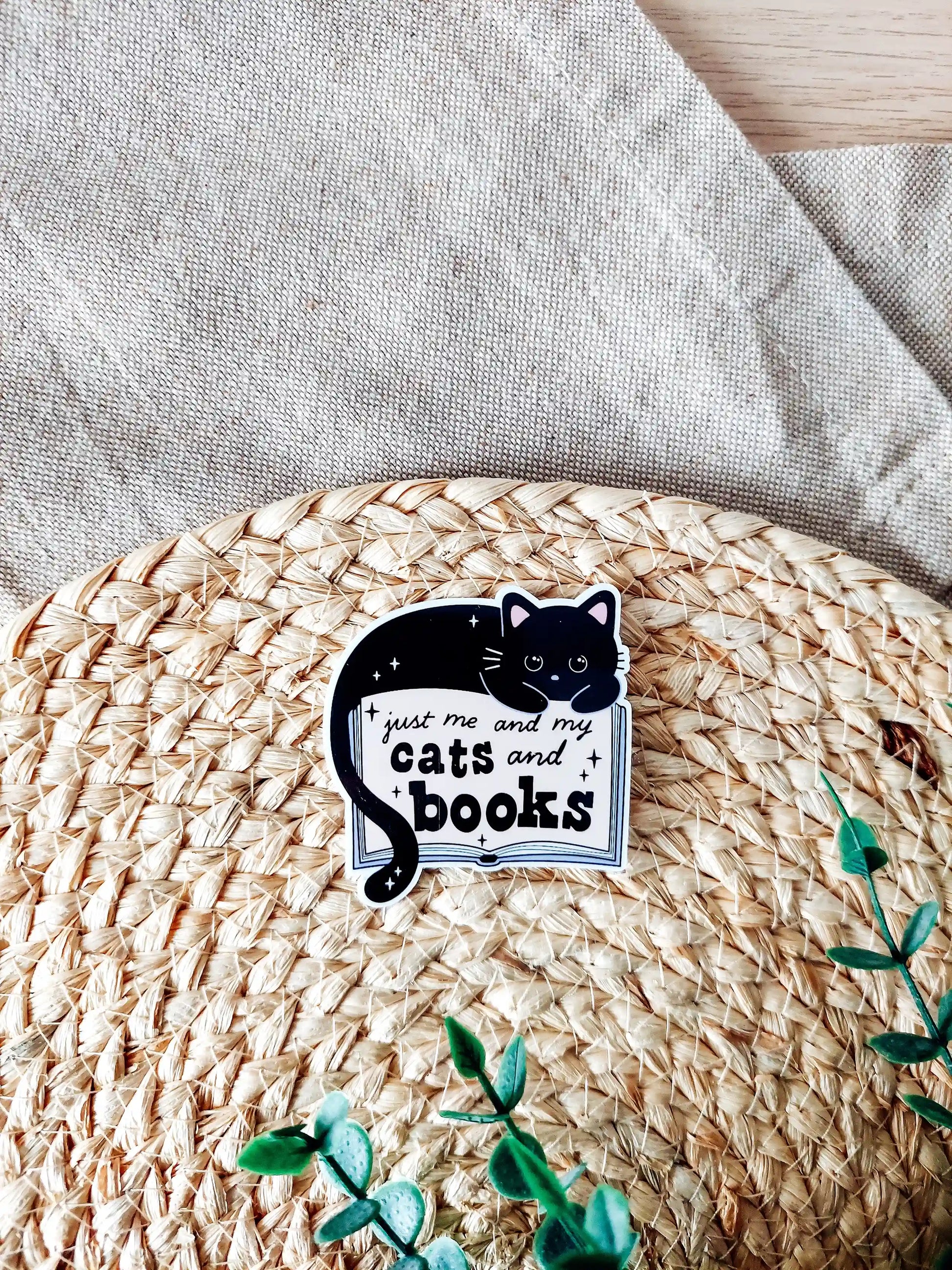 Vinyl sticker featuring a black cat lying on an open book with the text "Just me and my cats and books" photographed on a woven neutral background. 