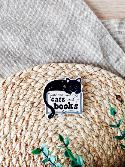 Vinyl sticker featuring a black cat lying on an open book with the text "Just me and my cats and books" photographed on a woven neutral background. 