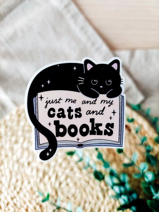 Vinyl sticker featuring a black cat lying on an open book with the text "Just me and my cats and books" photographed on a woven neutral background. 