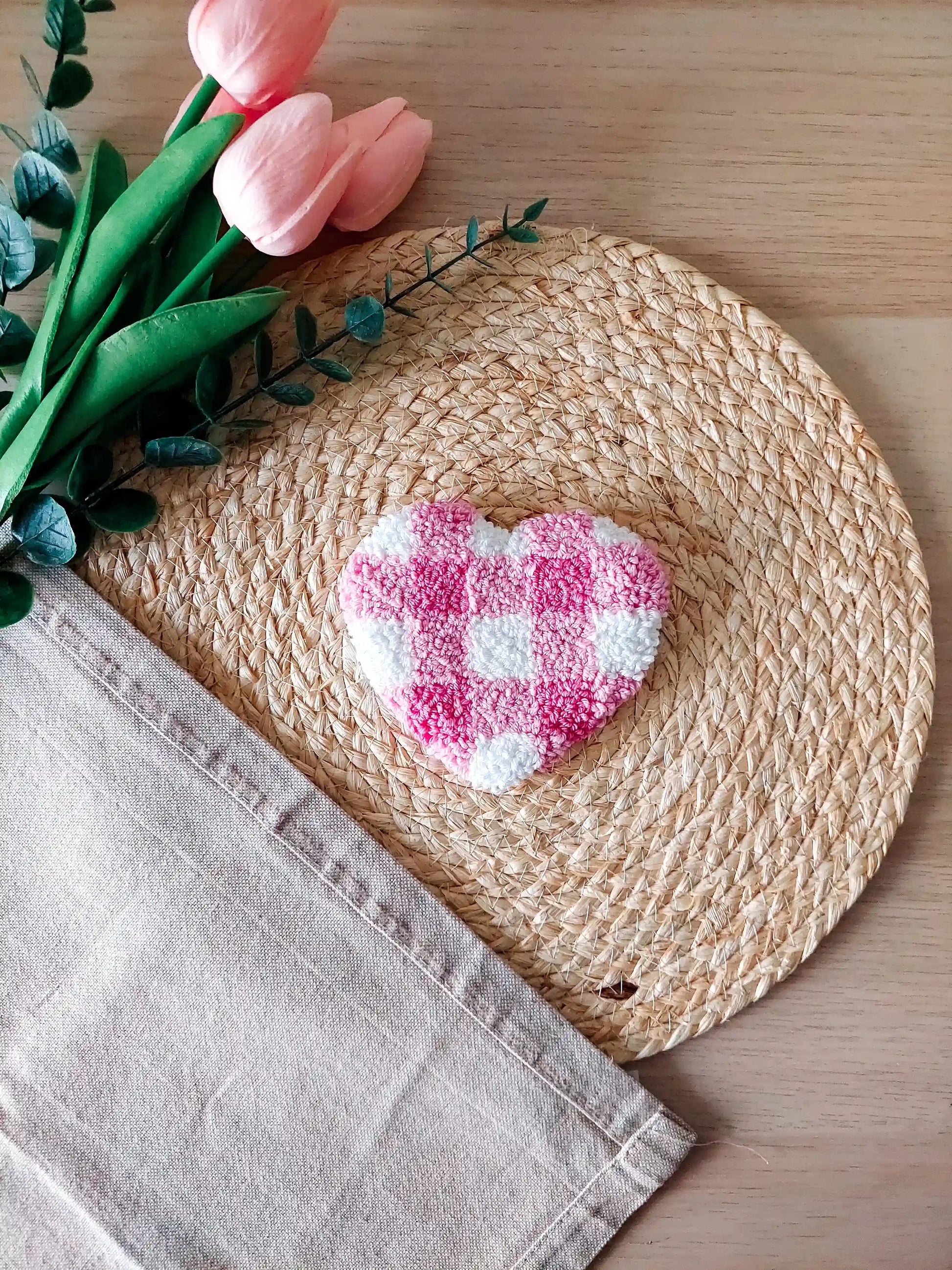 Handmade punch needle heart-shaped mug rug with a pink and white checkered pattern, photographed on a woven mat with decorative flowers in the back. 